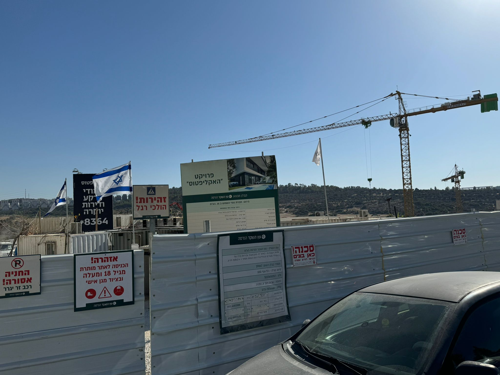 Construction site of the 'Eucalyptus' project in Beit Shemesh, showing cranes, safety signs, and Israeli flags against a clear sky. The site is surrounded by temporary fencing with various informational and warning signs in Hebrew, indicating active construction and safety regulations.