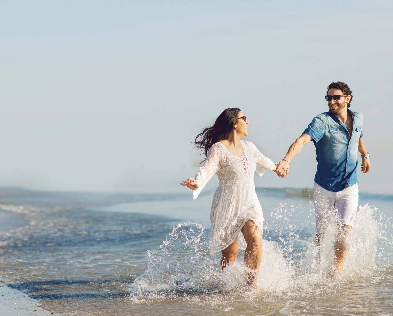 A couple enjoying a Mediterranean experience, holding hands and running along the shoreline with waves splashing around them. They are smiling and wearing casual beach attire, capturing a moment of joy and relaxation.