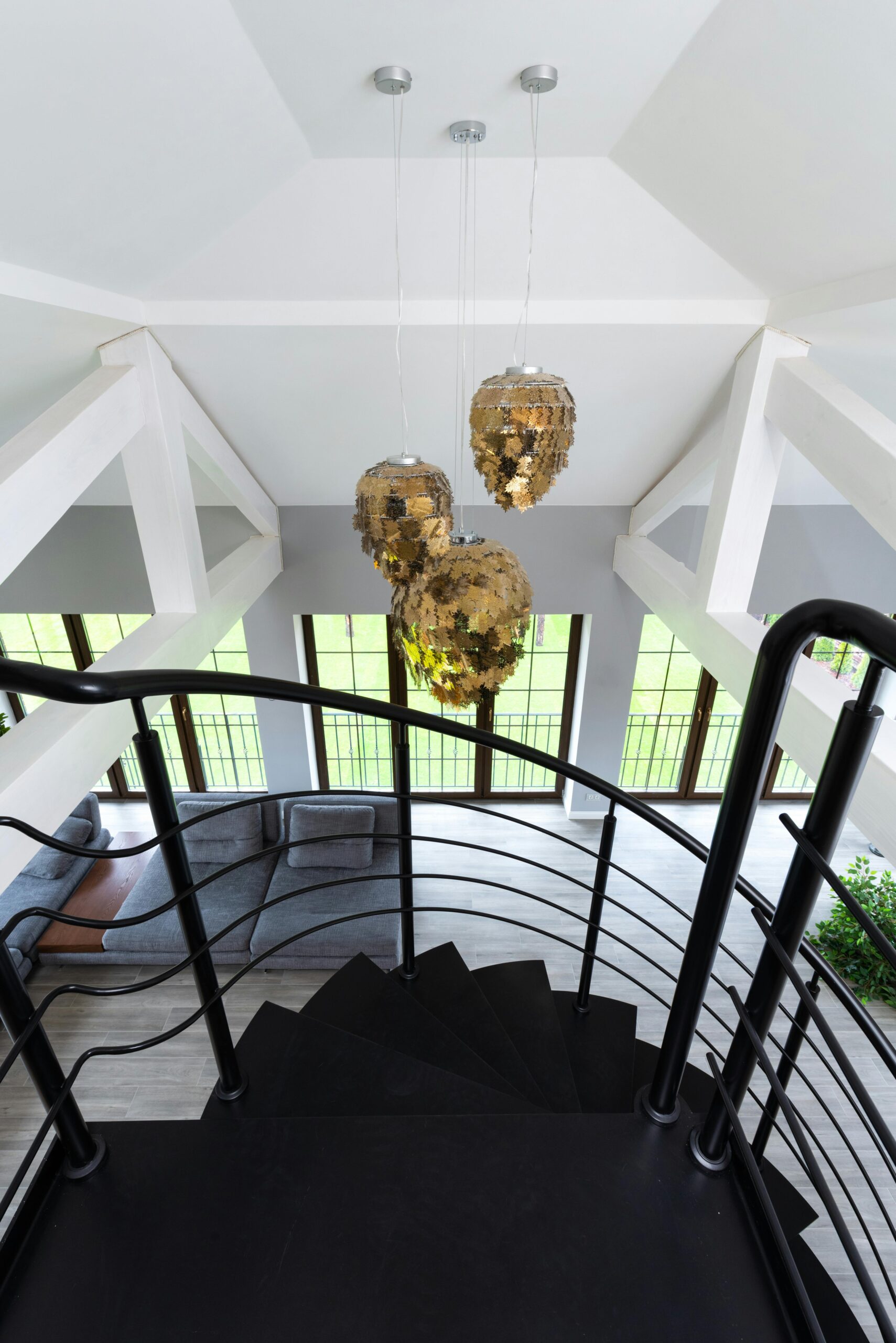 Interior view of a modern duplex featuring a spiral staircase with sleek black railings. The ceiling is vaulted with exposed white beams, and hanging from it are three ornate, golden pendant lights. The lower level has large windows with dark frames, allowing natural light to fill the space. A gray sectional sofa is arranged in the living area, with light wood flooring throughout the space. The design combines contemporary and elegant elements.