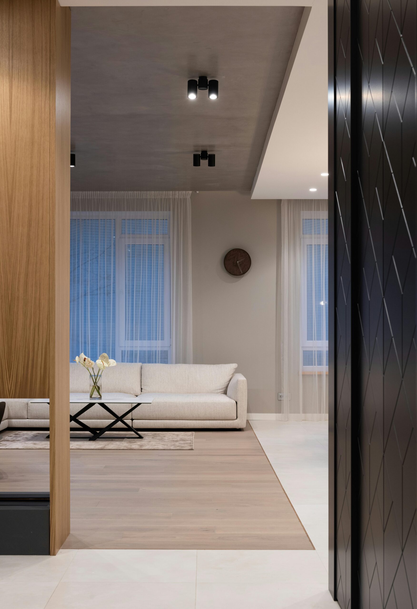 Stylish penthouse living area with a minimalist design. The space features a light beige sofa against a taupe wall, accented by sheer white curtains over large windows. A sleek black coffee table with a geometric base sits in front of the sofa, topped with a simple vase holding white flowers. The ceiling has a modern industrial touch with exposed concrete and recessed lighting fixtures. Warm wood paneling contrasts with darker textured walls, creating a refined and sophisticated ambiance.