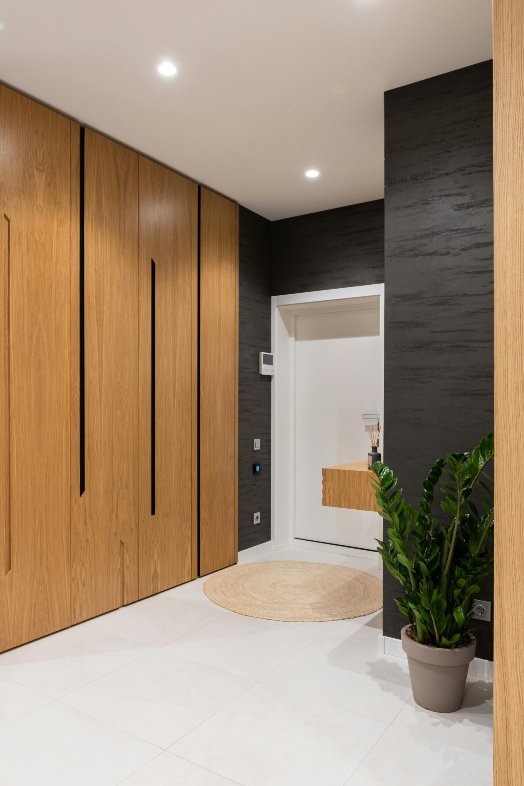 Modern apartment entryway with a sleek and minimalist design. The space features a mix of light wood and dark textured walls. On one side, there are vertical wood panels with integrated handles, likely concealing storage. The door is painted white with a small floating wooden shelf beside it, adorned with decorative items. The flooring is light-colored tile, complemented by a round woven rug. A potted green plant adds a touch of nature to the otherwise neutral-toned space, creating a warm and welcoming atmosphere.