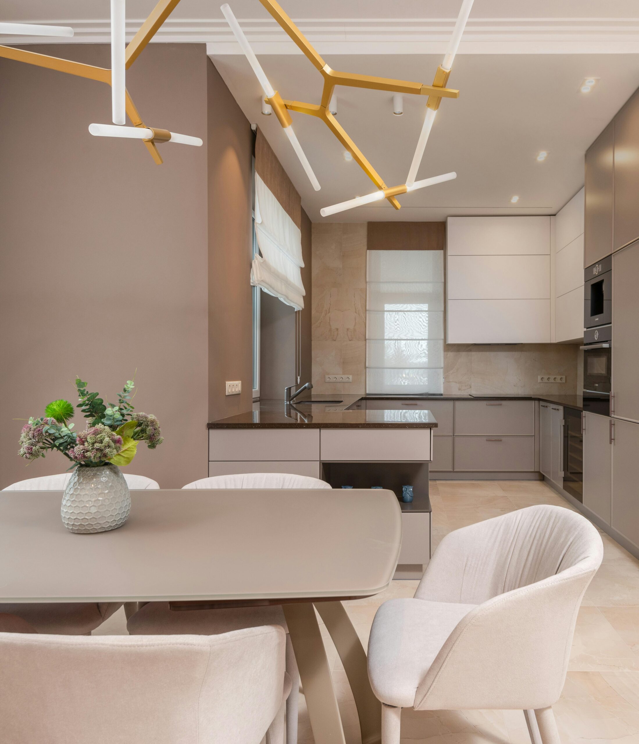 Modern apartment kitchen and dining area with a minimalist design. The kitchen features sleek cabinets in neutral tones, a built-in oven, and a glossy countertop. A beige dining table with matching upholstered chairs is decorated with a simple vase of flowers. Above the table hangs a contemporary light fixture with gold and white branches, adding a stylish touch to the space. The color scheme is warm and cohesive, creating a cozy yet elegant atmosphere.