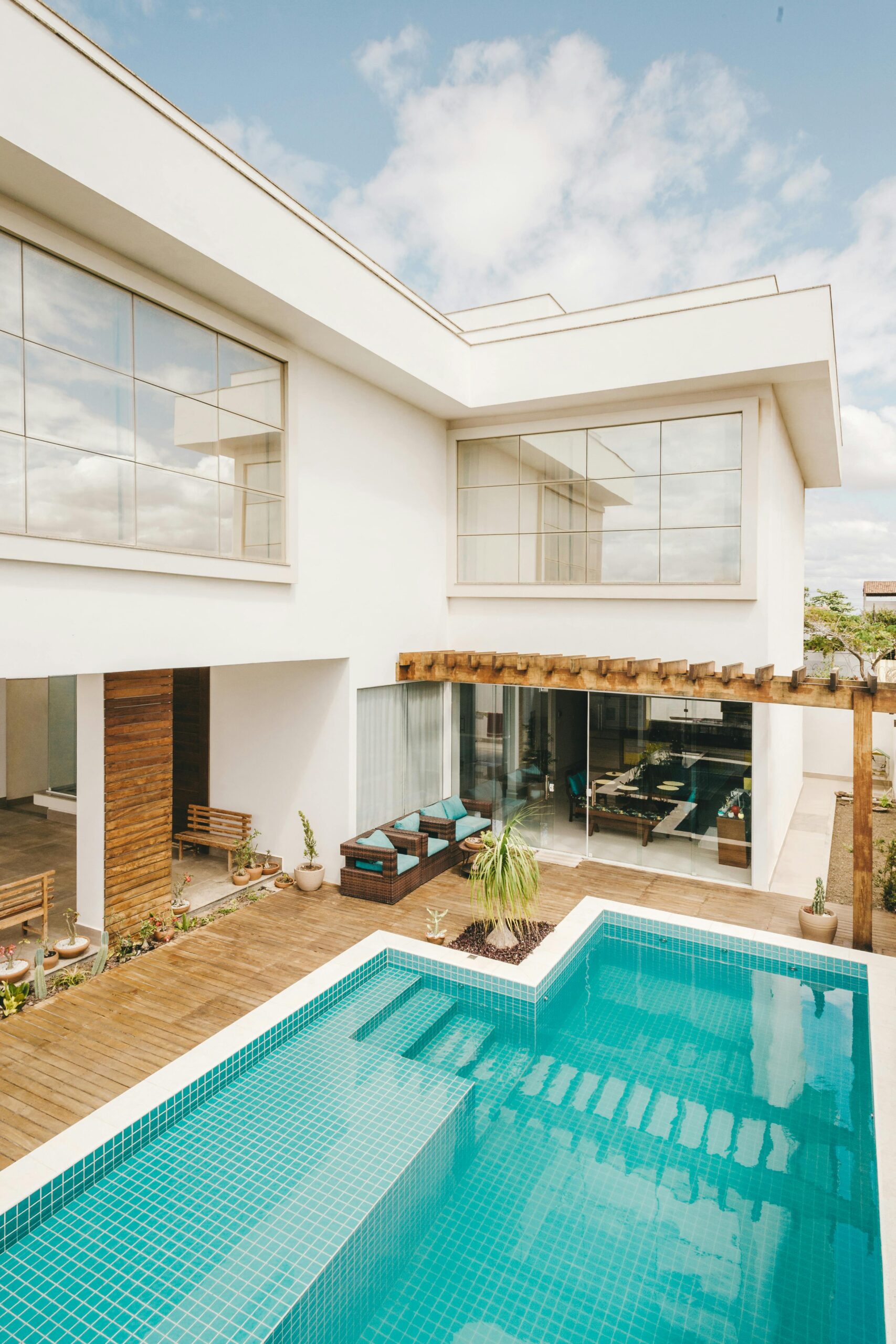 Modern two-story house with large glass windows and a minimalist design. The exterior features clean white walls, a wooden deck, and a blue-tiled swimming pool in the courtyard. The seating area under a wooden pergola includes rattan furniture with teal cushions, surrounded by potted plants. The overall style is contemporary and inviting.