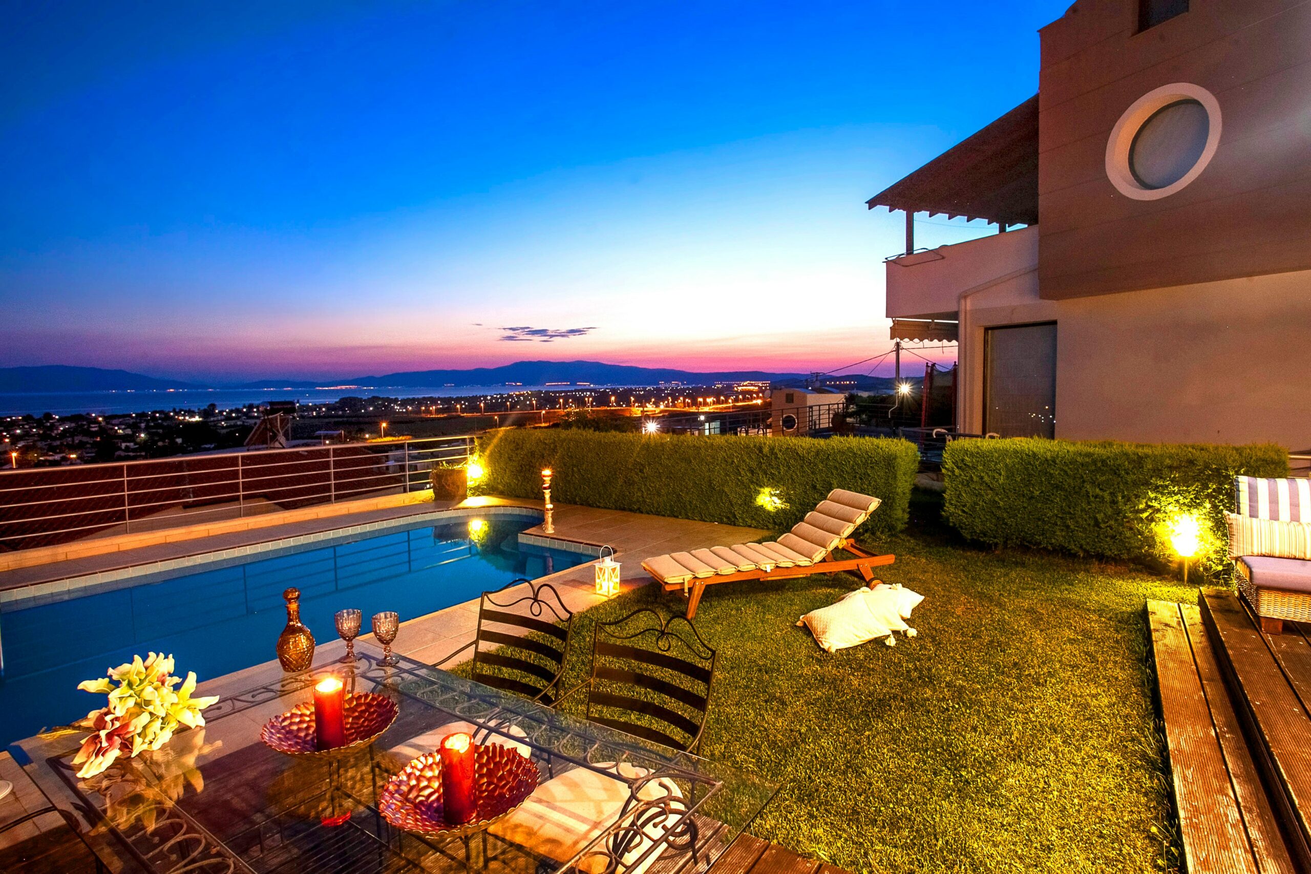 A luxurious vacation home with a stunning outdoor area at sunset. The scene features a private pool, surrounded by manicured greenery, and an inviting lounging area. The setup includes cozy lounge chairs, a stylish outdoor dining table, and ambient lighting, all overlooking a beautiful panoramic view of distant mountains and city lights. The setting is warm and serene, perfect for a relaxing getaway.