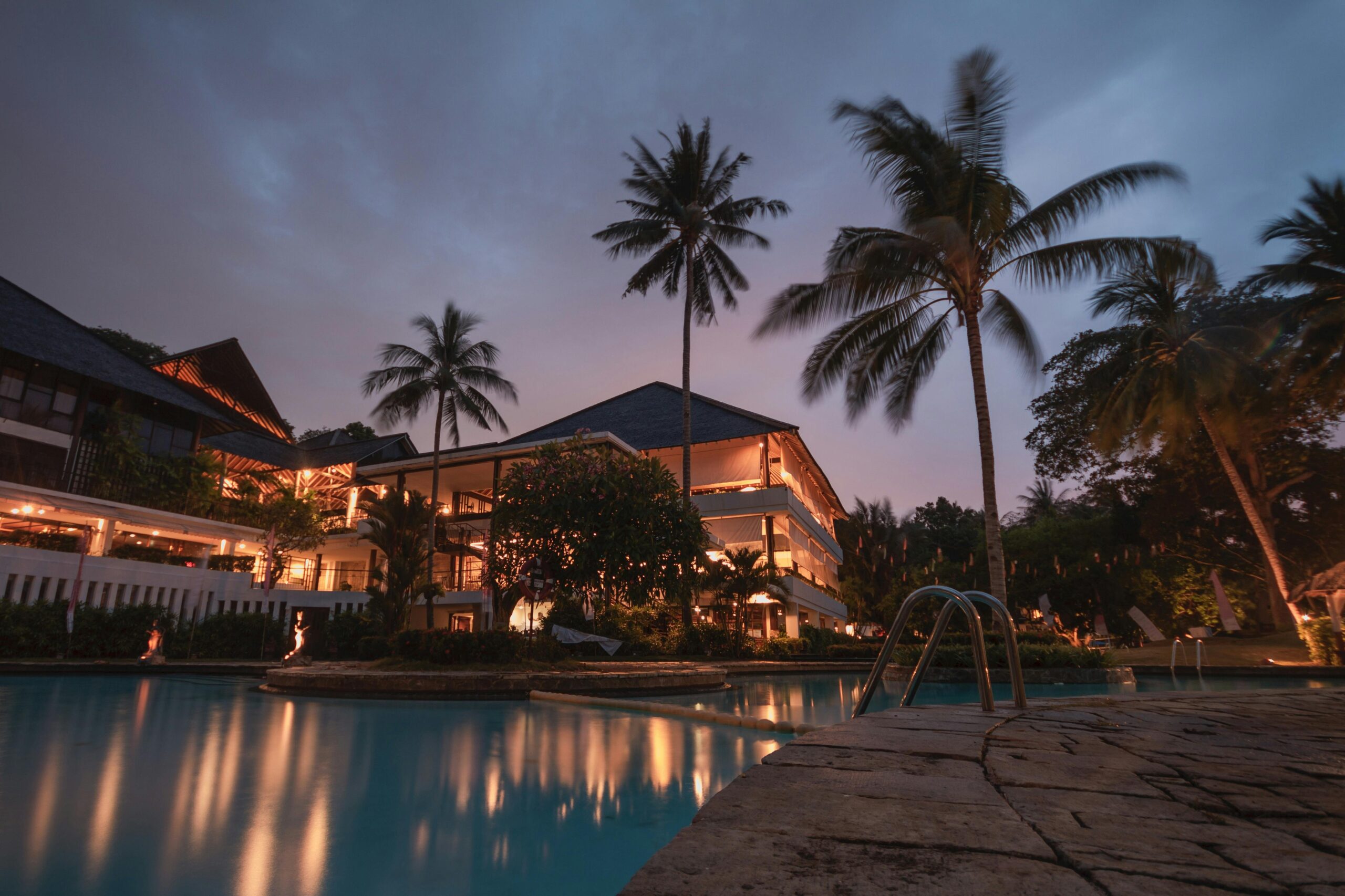 A luxurious tropical villa illuminated at dusk, surrounded by tall palm trees and lush greenery. The scene showcases a large swimming pool reflecting the warm lights of the villa’s exterior, creating a tranquil and inviting atmosphere. The multi-story villa features spacious balconies, warm ambient lighting, and an elegant, open design perfect for a tropical getaway. The calming blue of the pool contrasts beautifully with the darkening sky, offering an idyllic retreat setting.