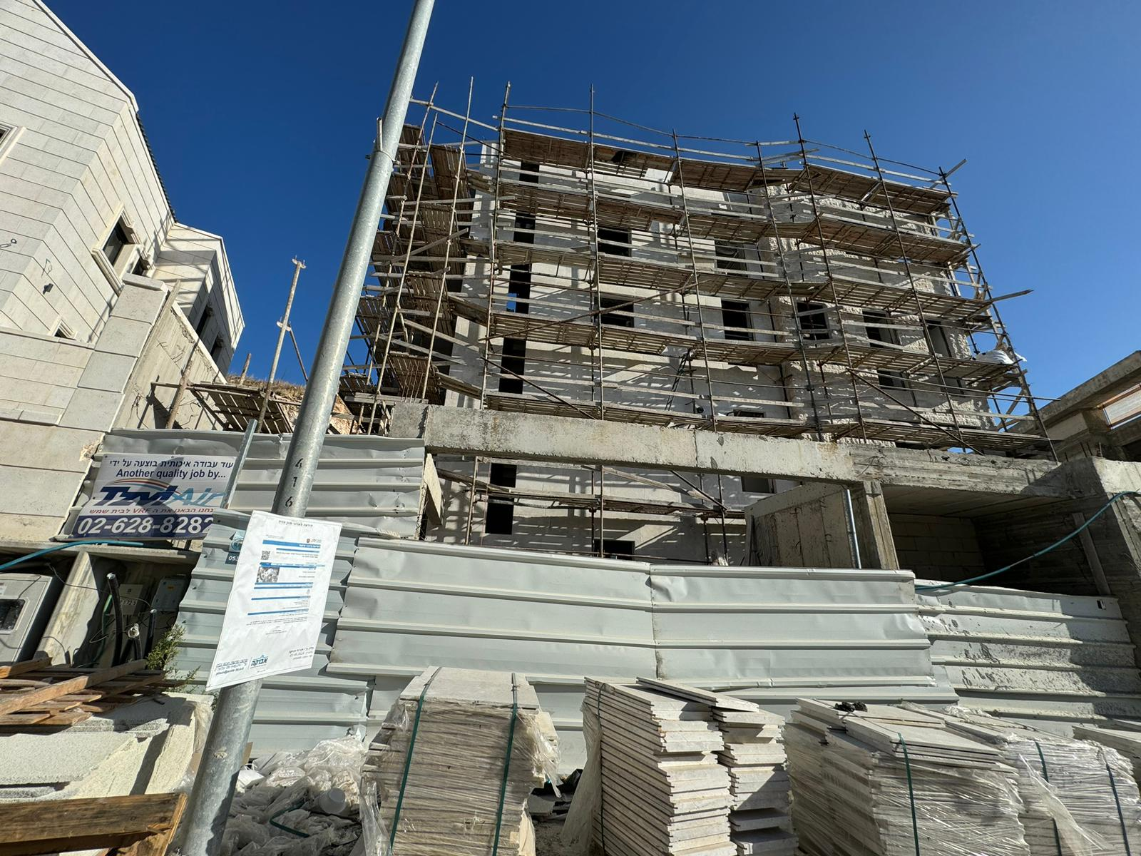 Construction site of a multi-story residential building in Ramat Beit Shemesh, with scaffolding surrounding the structure. The building is in the advanced stages of construction, and a development permit notice is displayed on a nearby pole. The site is marked with signage indicating ongoing high-quality work by a local construction company, with materials neatly stacked in the foreground.