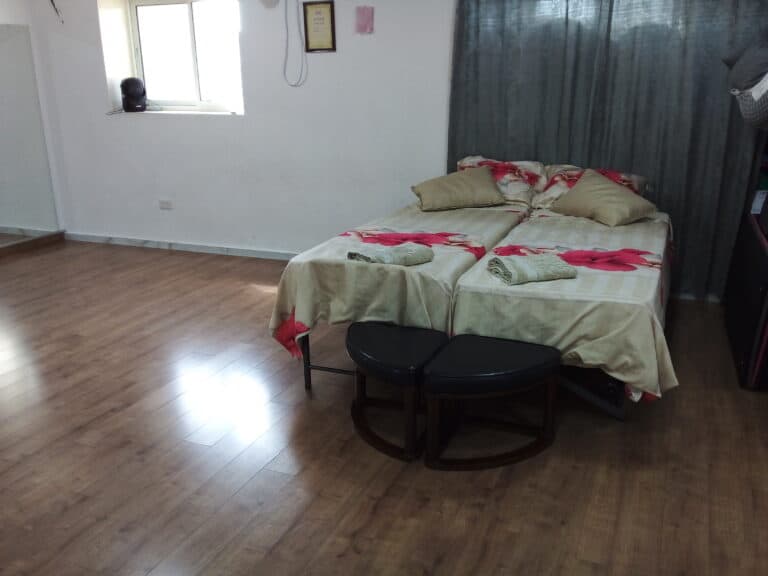 A bedroom with a bed, two pillows, and a black ottoman. The bed has a floral patterned comforter and a green blanket. The room has a wooden floor and a window with white curtains.
