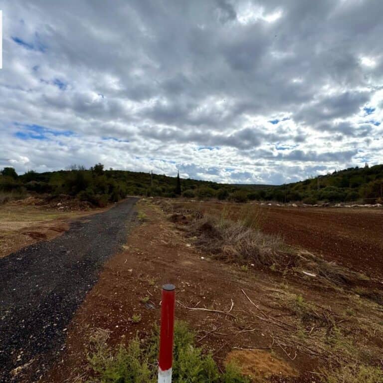 A dirt road winds through a rural landscape, flanked by lush green vegetation and a cloudy sky.