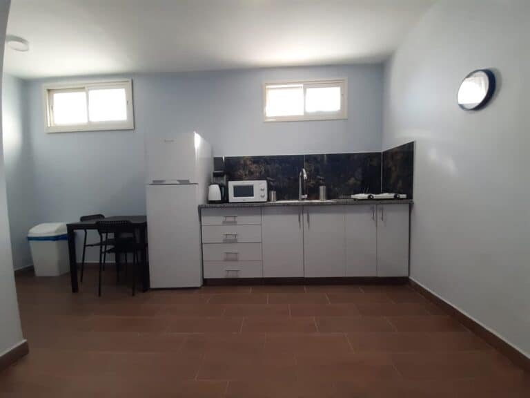 The image shows a small, modern kitchen area. The floor is covered with reddish-brown tiles, and the walls are painted in a light blue color. On the left side of the room, there is a white refriger...