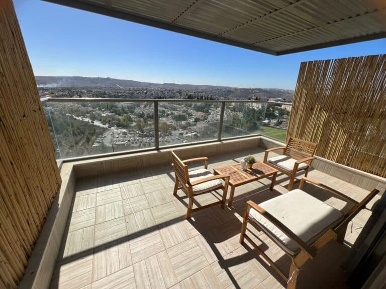 A balcony with a wooden table and chairs overlooking a cityscape, featuring a bamboo fence and a clear blue sky.