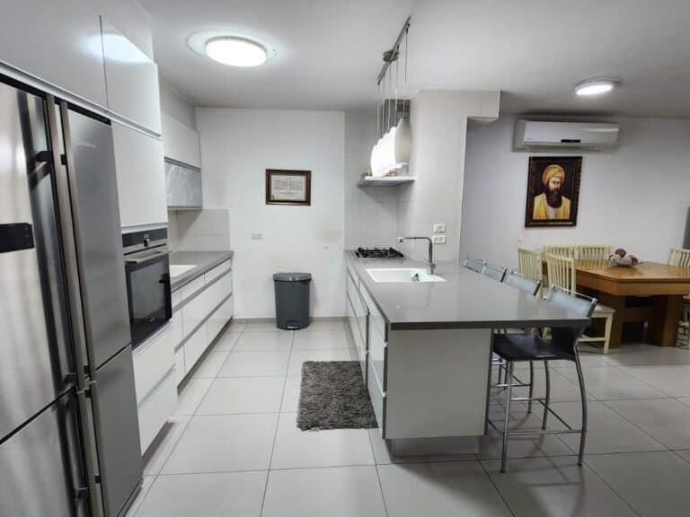 The image shows a modern kitchen with a clean, minimalist design. The kitchen features sleek, white cabinetry and countertops, creating a cohesive look. On the left side, there is a stainless steel...