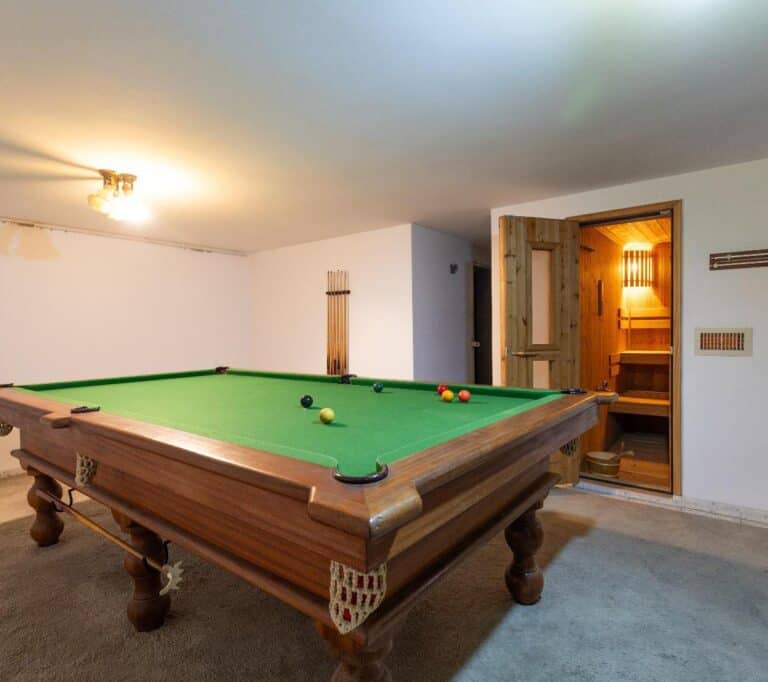 The image shows a cozy room with a pool table as the main focus. The pool table has a green felt surface and is surrounded by wooden legs. There are several pool balls on the table, including a bla...