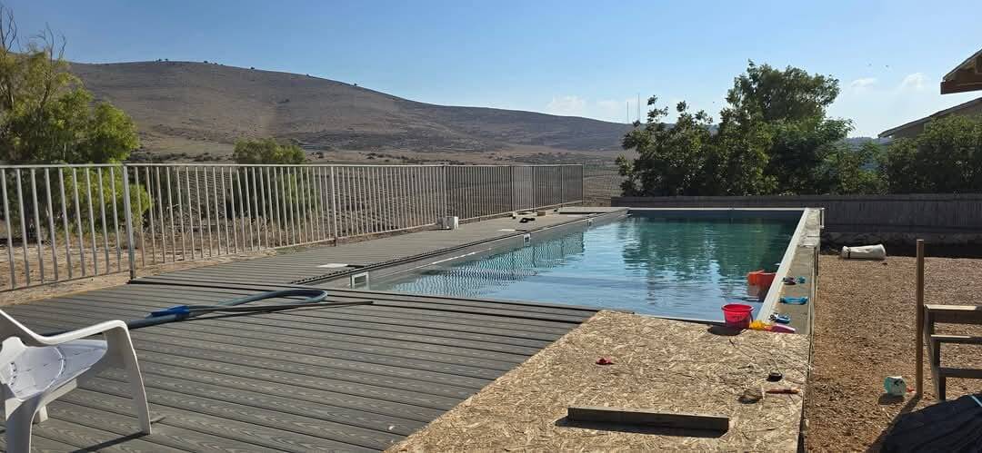 The image shows a large swimming pool surrounded by a wooden deck. The pool is rectangular and filled with clear blue water. On the left side of the pool, there is a white chair and a blue pool sho...