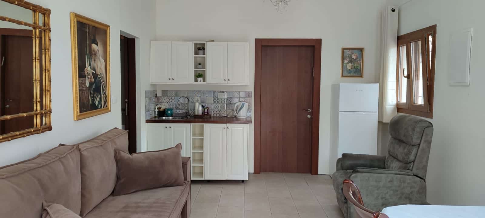 The image shows a cozy living room area that seamlessly transitions into a kitchenette. The living room features a beige sofa adorned with two brown throw pillows, positioned against a wall decorat...