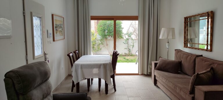The image shows a cozy living room with a dining area. The room features a beige sofa adorned with brown cushions and a matching throw blanket. A wooden floor lamp with a white shade stands to the...