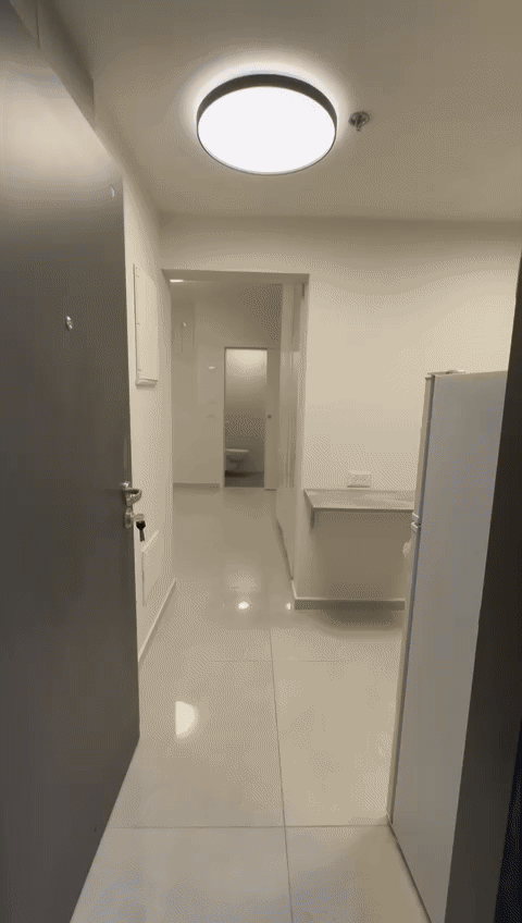 The image shows a modern hallway with white walls and tiled floors. The ceiling features recessed lighting that casts a bright, even light throughout the space. On the left side of the hallway, the...