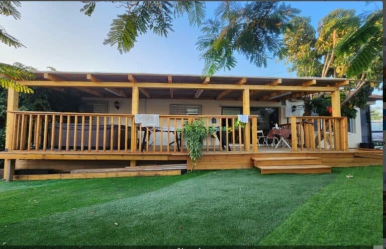 The image shows a backyard with a wooden deck area. The deck has a railing and is supported by wooden beams. There is a table and chairs set up on the deck, suggesting it's a space for outdoor dini...