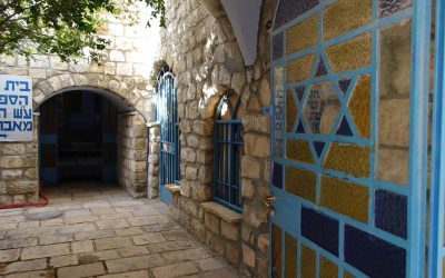 A stone building with arched windows and a blue door, featuring a Star of David design, with a tree and a sign in Hebrew script visible in the background.