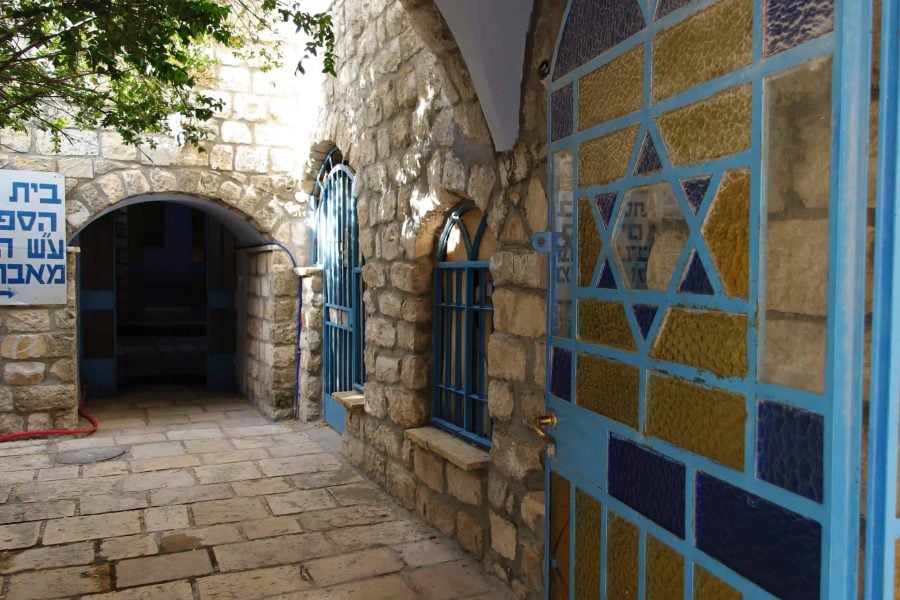 A stone building with arched windows and a blue door, featuring a Star of David design, with a tree and a sign in Hebrew script visible in the background.