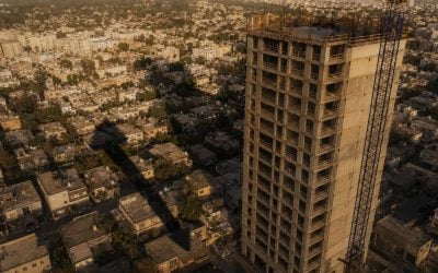 A high overhead shot at golden hour of a half-built raw-concrete apartment tower rising over a block of small 1950s low-rise homes in Tel Aviv. A single tall construction crane throws a long shadow across the old rooftops, and the shadow happens to point upward like an arrow. Warm low-angle light, faint dust haze in the air, deep shadows, cinematic, shot on a 35mm lens, strong sense of scale between the giant new tower and the tiny old houses below. Editorial and conceptual, not a stock photo. No text, no words, no numbers, no logos anywhere in the image.