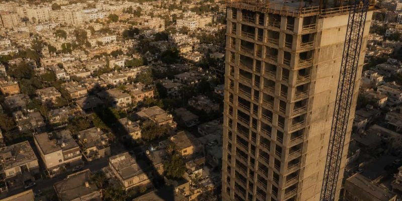 A high overhead shot at golden hour of a half-built raw-concrete apartment tower rising over a block of small 1950s low-rise homes in Tel Aviv. A single tall construction crane throws a long shadow across the old rooftops, and the shadow happens to point upward like an arrow. Warm low-angle light, faint dust haze in the air, deep shadows, cinematic, shot on a 35mm lens, strong sense of scale between the giant new tower and the tiny old houses below. Editorial and conceptual, not a stock photo. No text, no words, no numbers, no logos anywhere in the image.