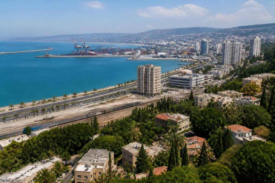 Aerial view of Haifa Bay waterfront with port cranes, rail lines, and residential neighborhoods in daylight