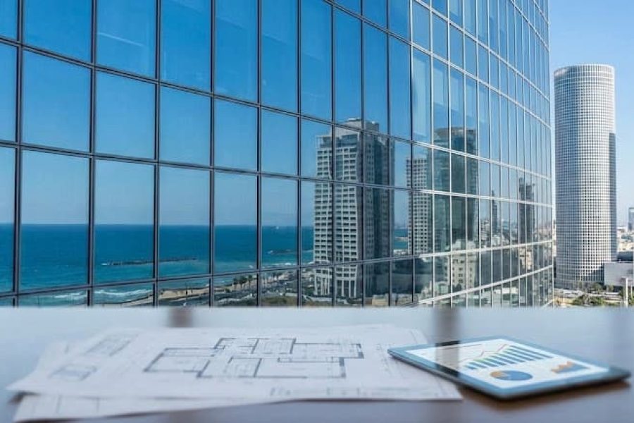 The image shows a modern office setting with large glass windows offering a view of a city skyline and the ocean. In the foreground, there is a desk with various items including architectural bluep...
